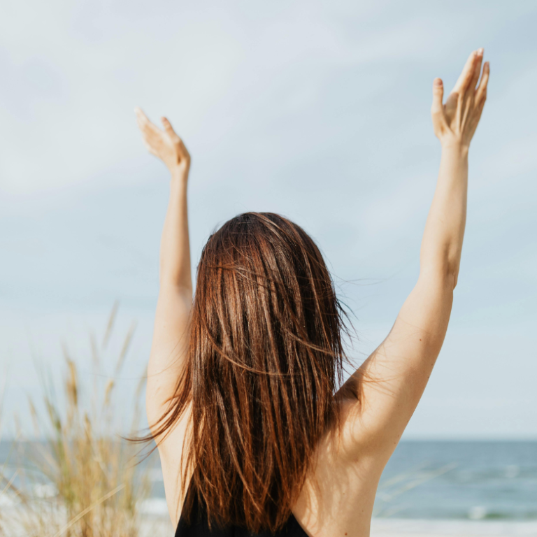 Frau mit erhobenen Händen steht am Strand vor dem Meer und dem Himmel.