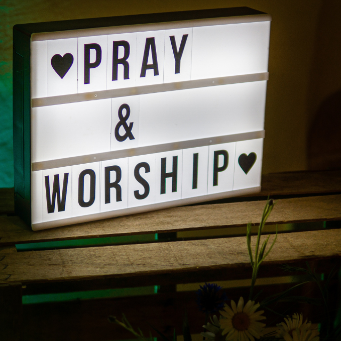 Leuchtkasten mit den Worten "PRAY & WORSHIP" auf einem Holzregal.